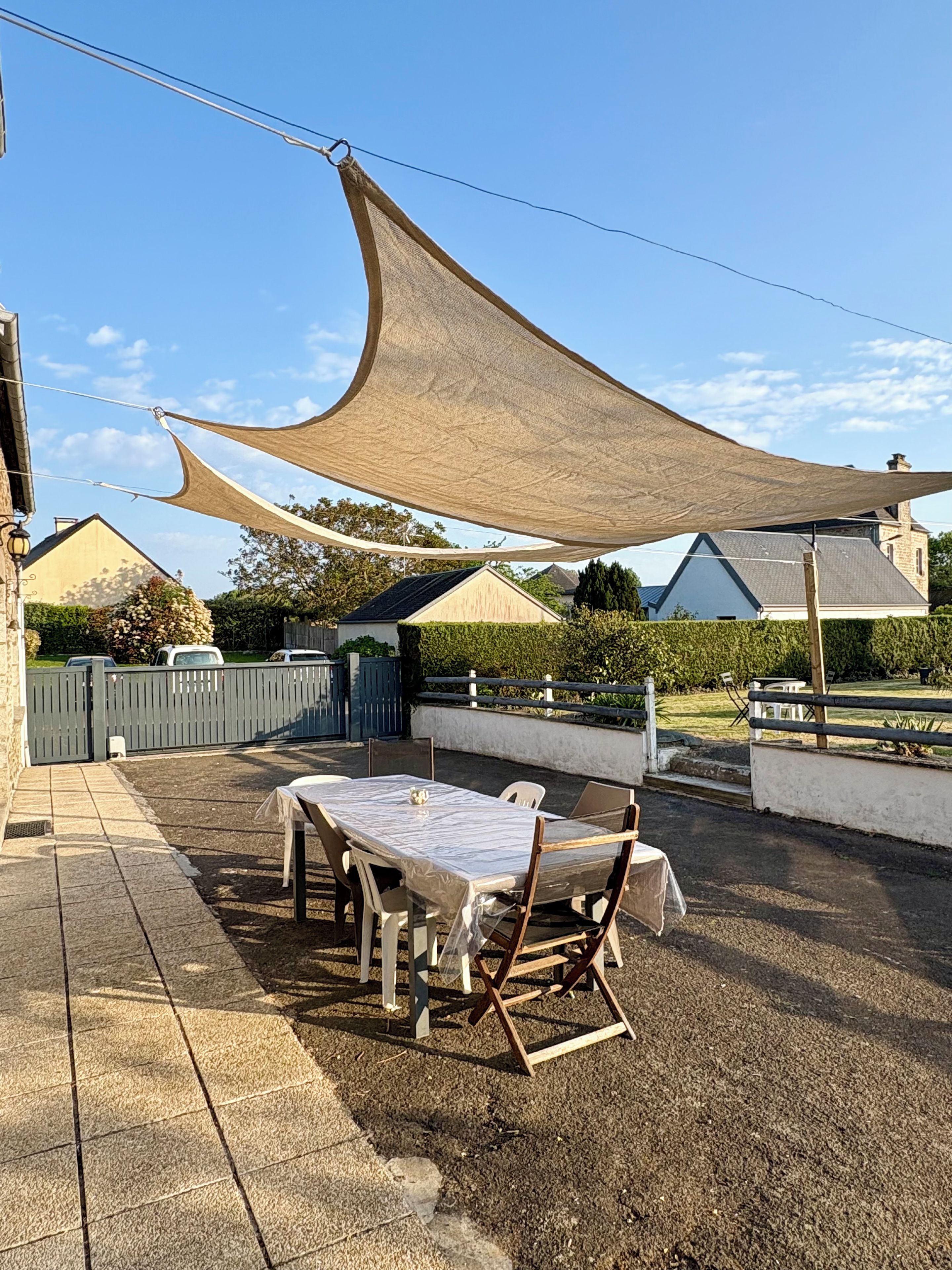 Une terrasse extérieure avec une table et des chaises, une grande voile d’ombrage et une vue sur les maisons au loin.
