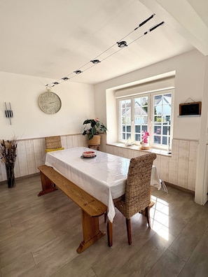 A dining area with a wooden table, bench, wicker chairs, a potted plant, and a window with a view. - My Little Farmhouse: A Quiet Family Home with an Enclosed Garden - 1.5 km from the sea 🌊 (Dragey-Ronthon)