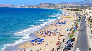A coastal cityscape with a beach, a road lined with parked cars, and buildings in the background. - Asikiko Suites - Rethymno Old Town Balcony Suite (Rethymno)