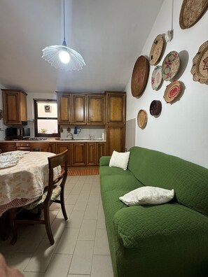 A kitchen with wooden cabinets, a dining table with chairs, a green sofa, and decorative woven baskets on the wall. - Domu 'e ajiajius Vacation Home (Gonnesa)