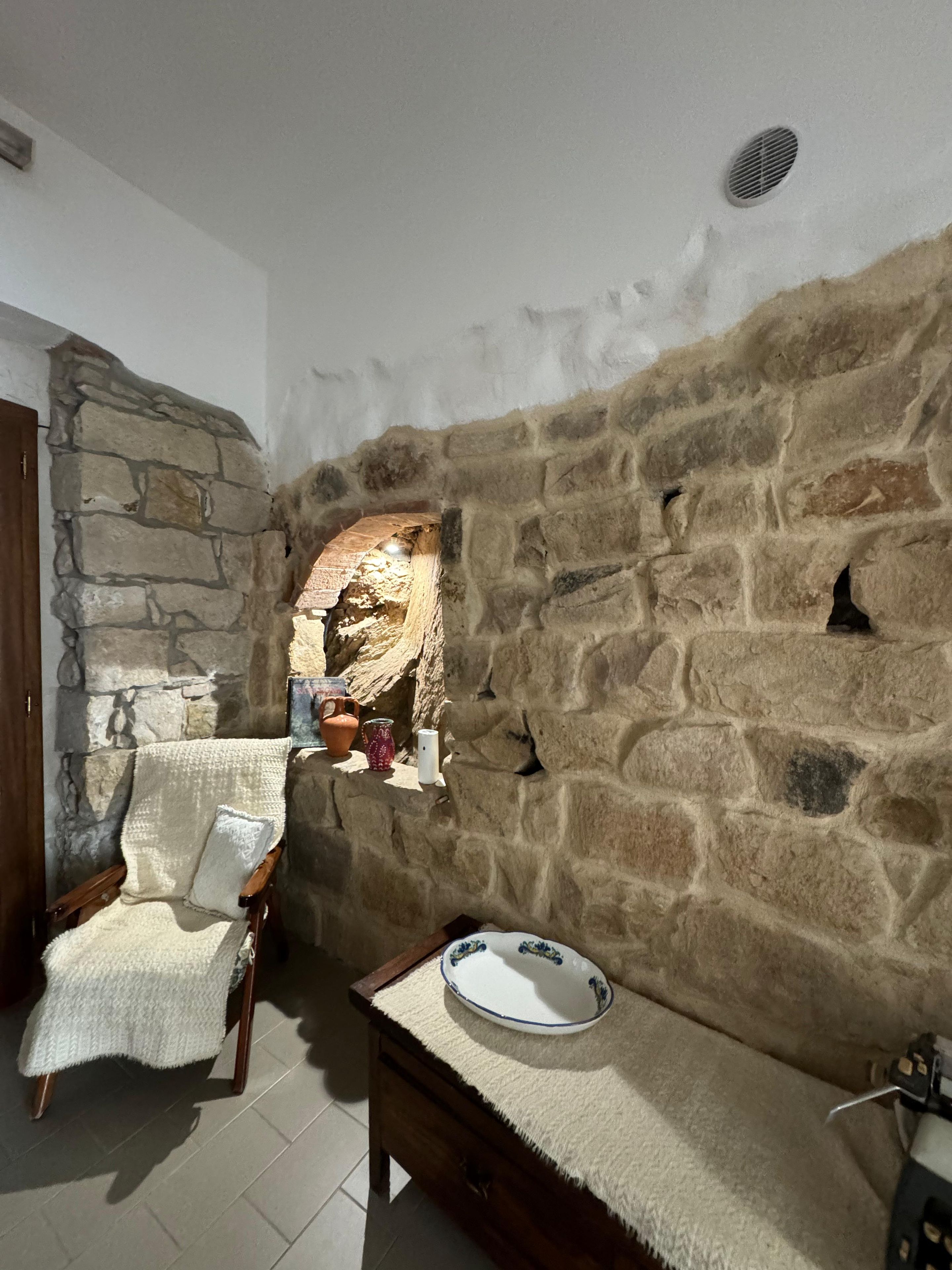 Une pièce aux murs de pierre, avec une chaise en bois, une petite table sur laquelle se trouve un bol, et une étagère ornée d’objets décoratifs.
