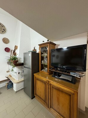 A kitchen with a refrigerator, a wooden cabinet with a glass display, and a television on a stand. - Domu 'e ajiajius Vacation Home (Gonnesa)