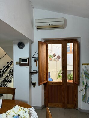 A room with a wooden door, an air conditioning unit, a clock, and a small window with a view of the outside. - Domu 'e ajiajius Vacation Home (Gonnesa)