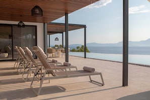 A sunlit outdoor seating area with lounge chairs and tables, overlooking a body of water and distant mountains.
