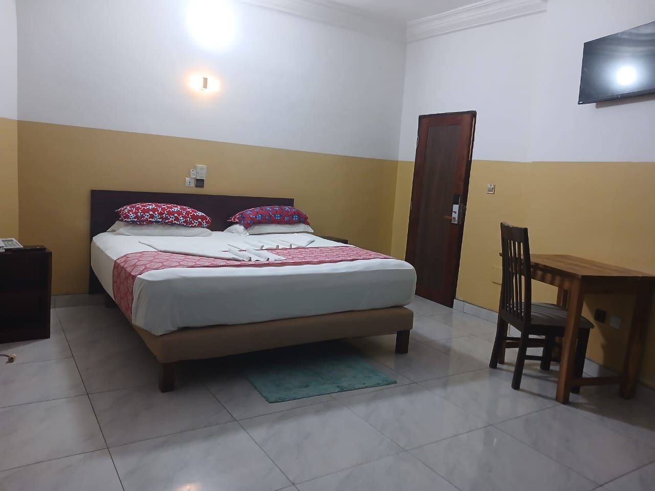 A bedroom with a bed, bedside table, chair, and a television on the wall.