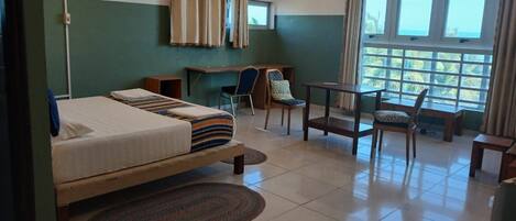 A hotel room with a bed, a desk, and a chair. There is a window with curtains and a view of palm trees.