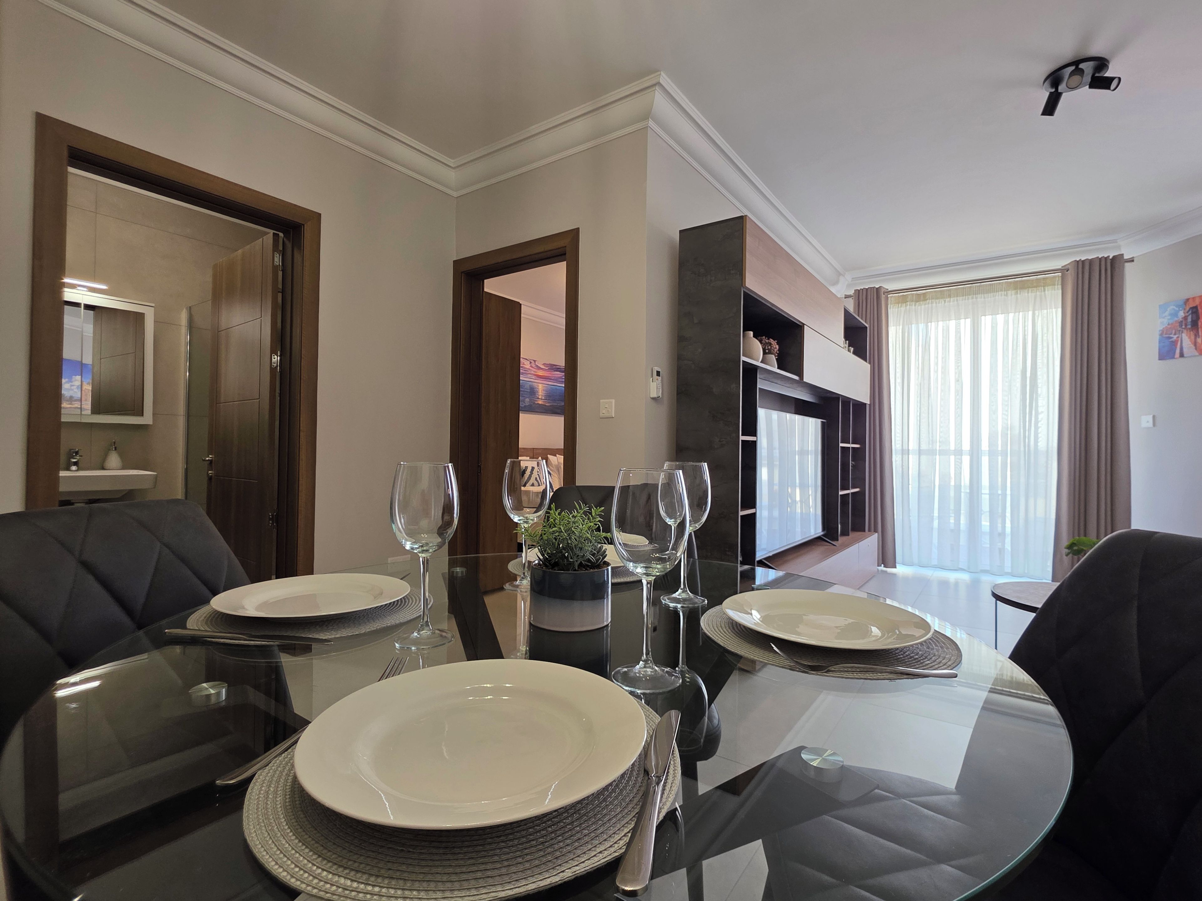 A dining area with a glass table set for two, a sofa, and a view of a balcony with curtains.
