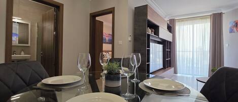 A dining area with a glass table set for two, a sofa, and a view of a balcony with curtains.