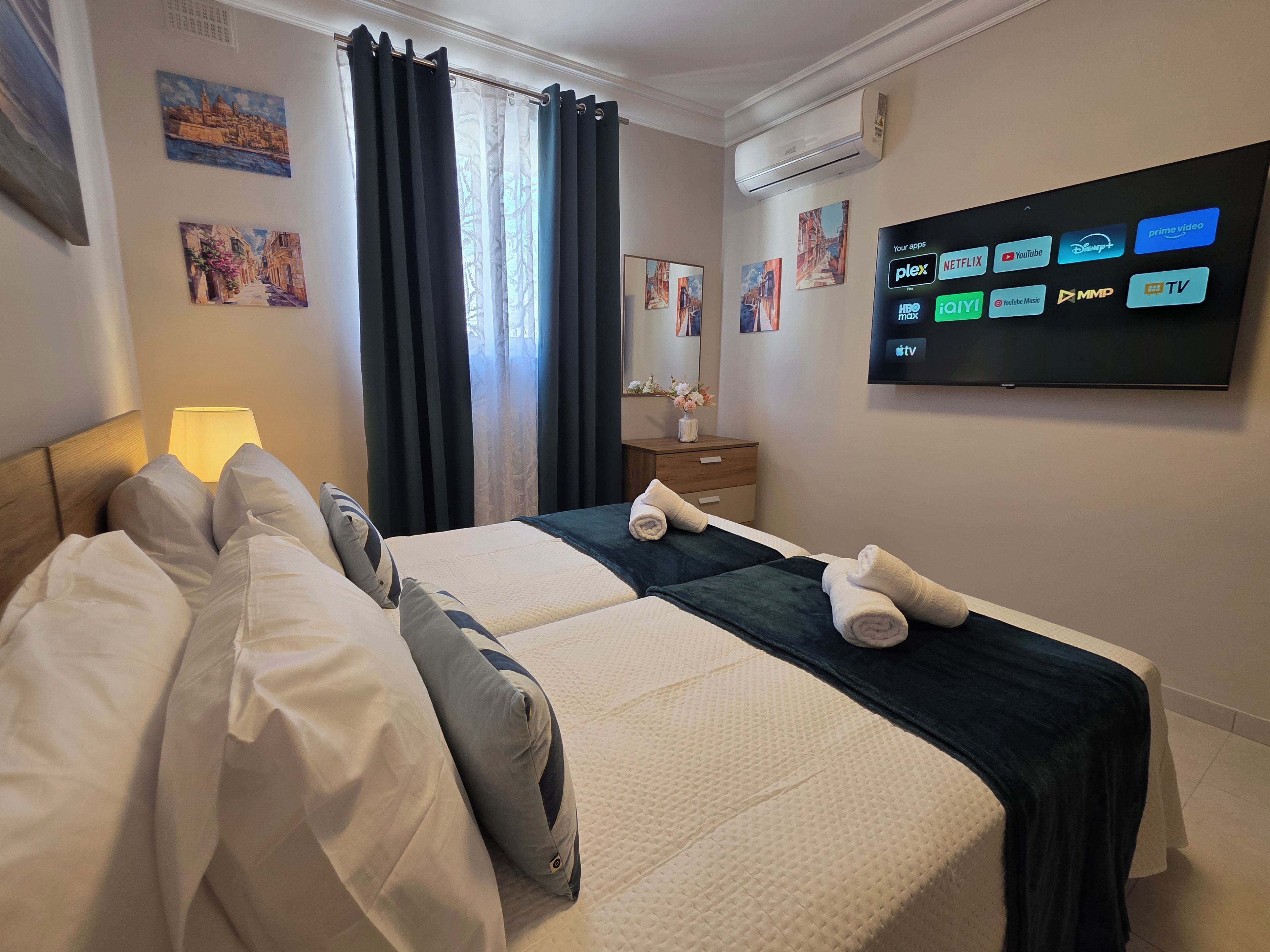 A hotel room with a bed, a TV displaying various streaming apps, a lamp, and artwork on the walls.