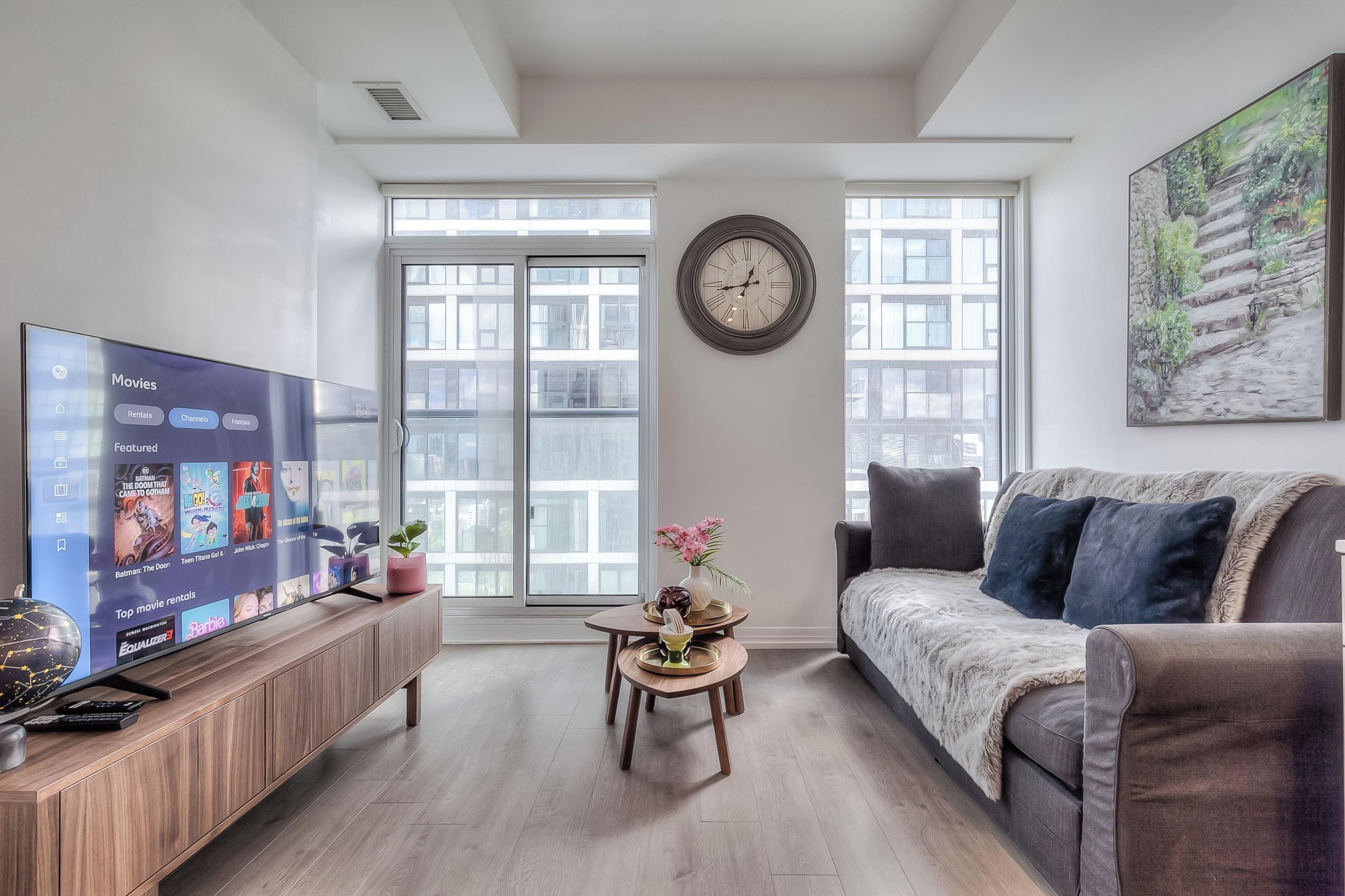 Una sala de estar con una televisión de pantalla plana mostrando opciones de películas, un reloj de pared redondo, un sofá, una mesa de centro con objetos decorativos y ventanas amplias con vistas a edificios.