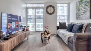 A living room with a flat-screen TV displaying movie options, a round wall clock, a sofa, a coffee table with decorative items, and large windows with a view of buildings.