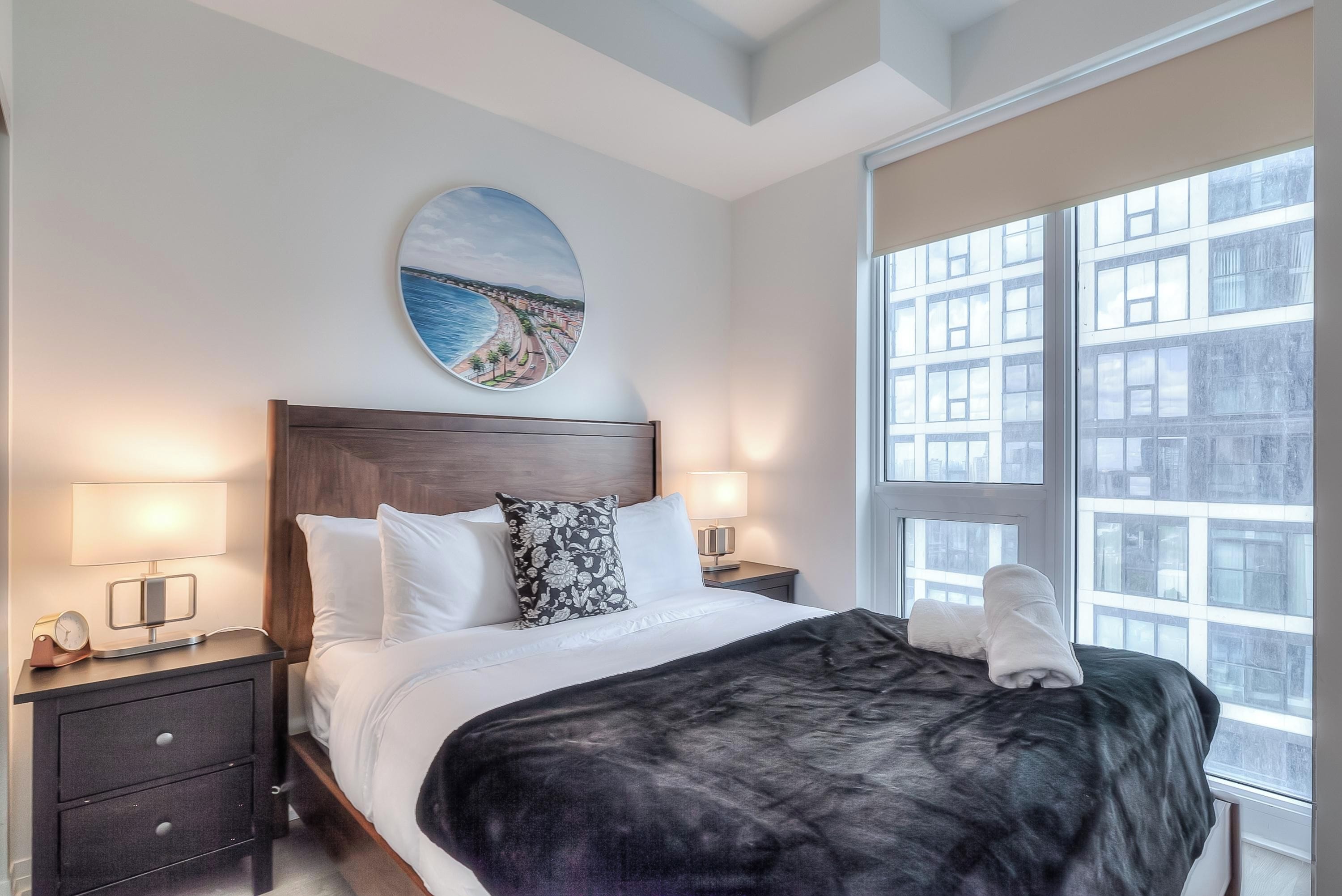 Habitación de hotel con una cama grande, dos mesitas de noche con lámparas, un cuadro redondo en la pared y vistas de edificios a través de la ventana.
