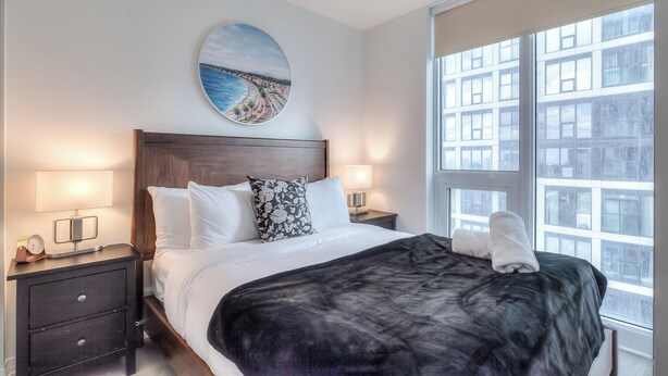 A hotel room with a large bed, two bedside tables with lamps, a round painting on the wall, and a view of buildings through the window.