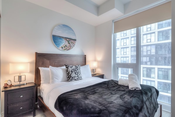 Une chambre d’hôtel avec un grand lit, deux tables de chevet avec des lampes, un tableau rond accroché au mur et une vue sur les bâtiments par la fenêtre.