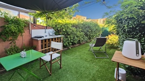 Een klein terras buiten met een groene tafel, twee stoelen, een klein barretje en een witte speaker.