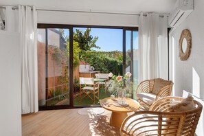 Una habitación moderna con puerta de vidrio corredera que da a un jardín, una mesa redonda de madera con sillas y un sofá de mimbre.