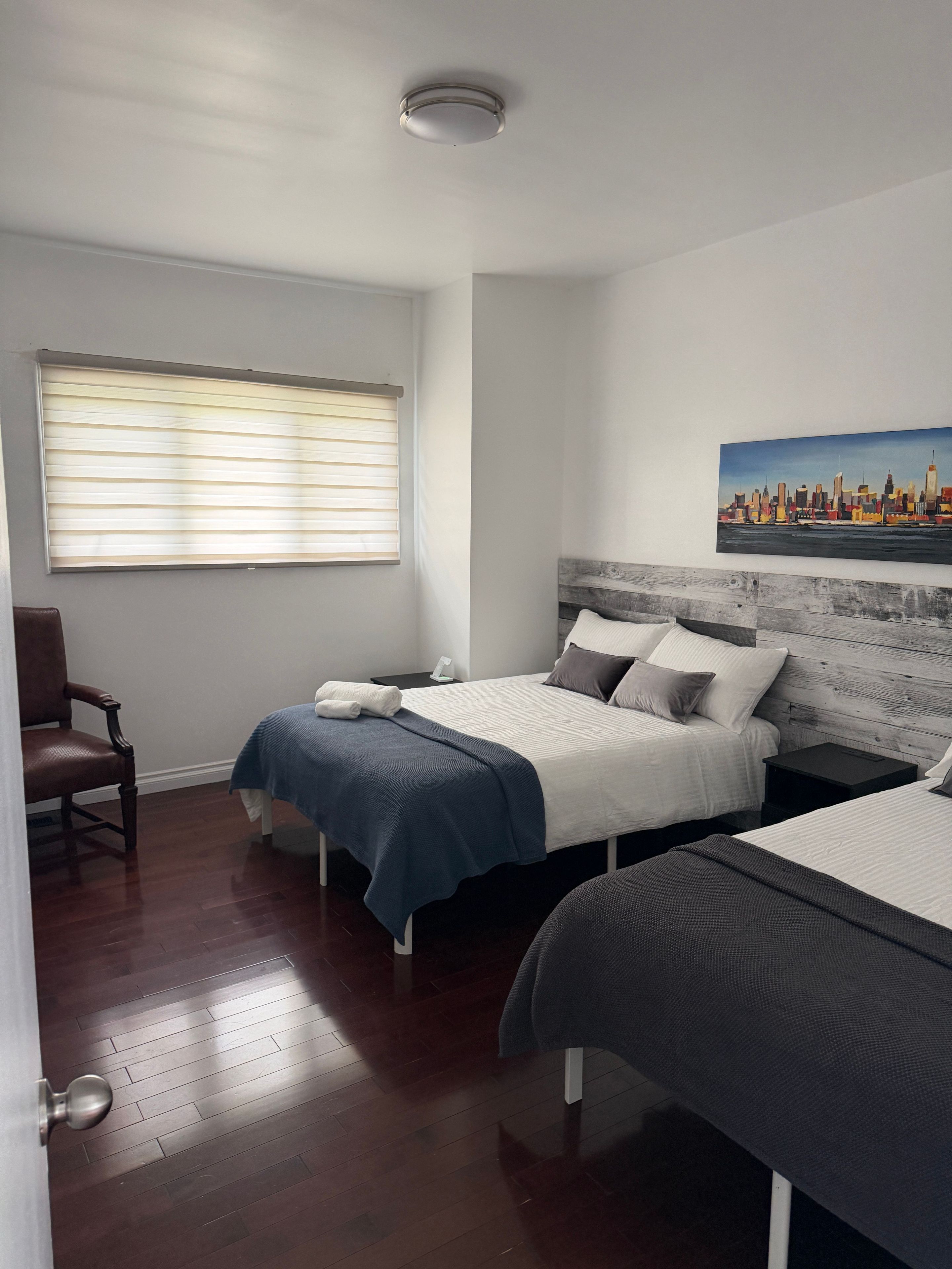 A bedroom with two beds, a chair, a painting, and a window with blinds.