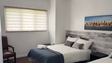 A bedroom with two beds, a chair, a painting, and a window with blinds.