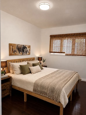 Un dormitorio con una cama de madera, una mesita de noche con lámpara, un cuadro en la pared y una ventana con persianas.
