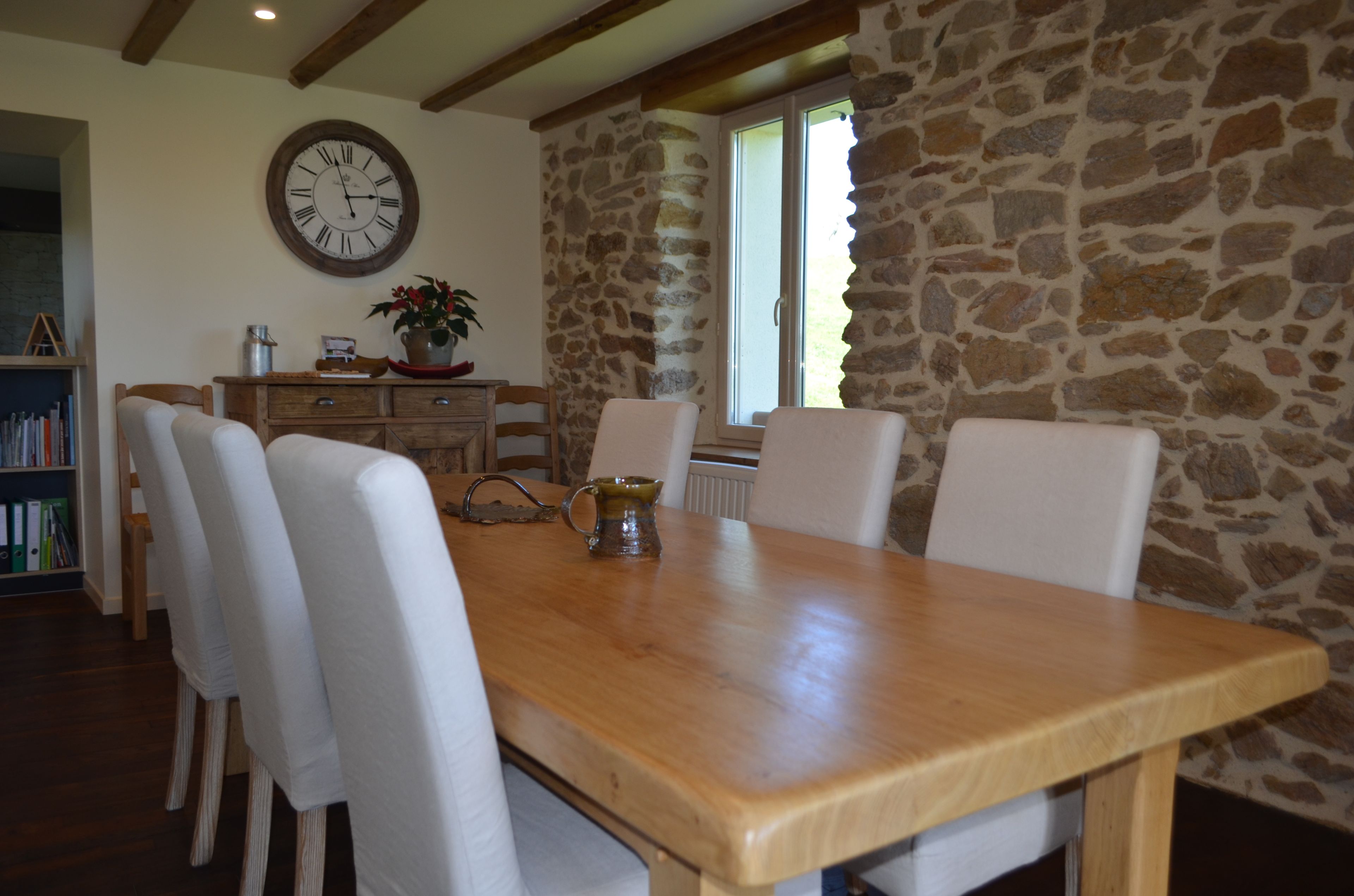 A dining area with a wooden table, white chairs, a clock on the wall, and a stone wall.