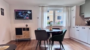 Un saló modern amb televisió plana penjada a la paret, una taula de dinar amb cadirees, un moble petit amb calaixos i una finestra amb cortines.