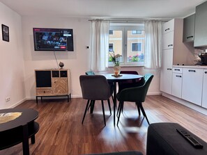 Ein modernes Wohnzimmer mit einem flachbildfernseher an der Wand, einem Esstisch mit Stühlen, einer kleinen Kommode und einem Fenster mit Vorhängen.