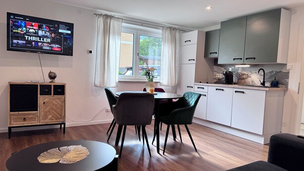 Un espai de vida modern amb televisió de pantalla plana, zona de menjador amb taula rodona i cadires, cuina amb armaris blancs i una finestra gran amb cortines.