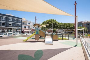 An outdoor playground with slides and climbing structures, surrounded by a paved area and buildings in the background. - Home2Book Betty Modern Studio in Puerto (Puerto del Rosario)