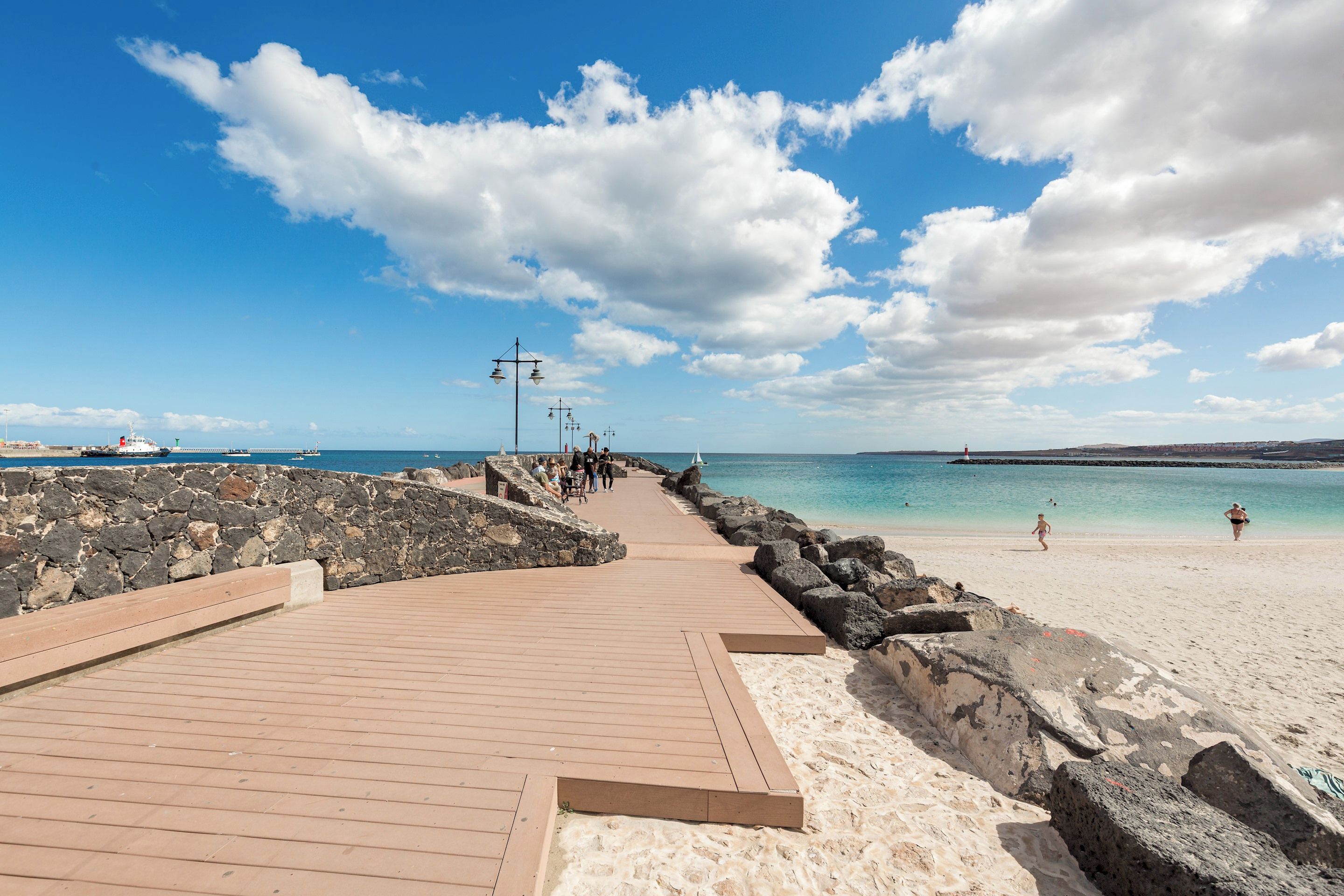 Eine Holzpromenade führt zu einem Sandstrand, der von einem klaren Himmel mit vereinzelten Wolken überblickt.
