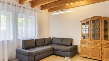 Ein modernes Wohnzimmer mit einem Ecksofa, einer Holzkommode und großen Fenstern mit durchsichtigen Vorhängen.