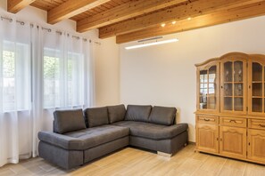 Een moderne woonkamer met een hoekbank, een houten kast en grote ramen met lichte gordijnen.