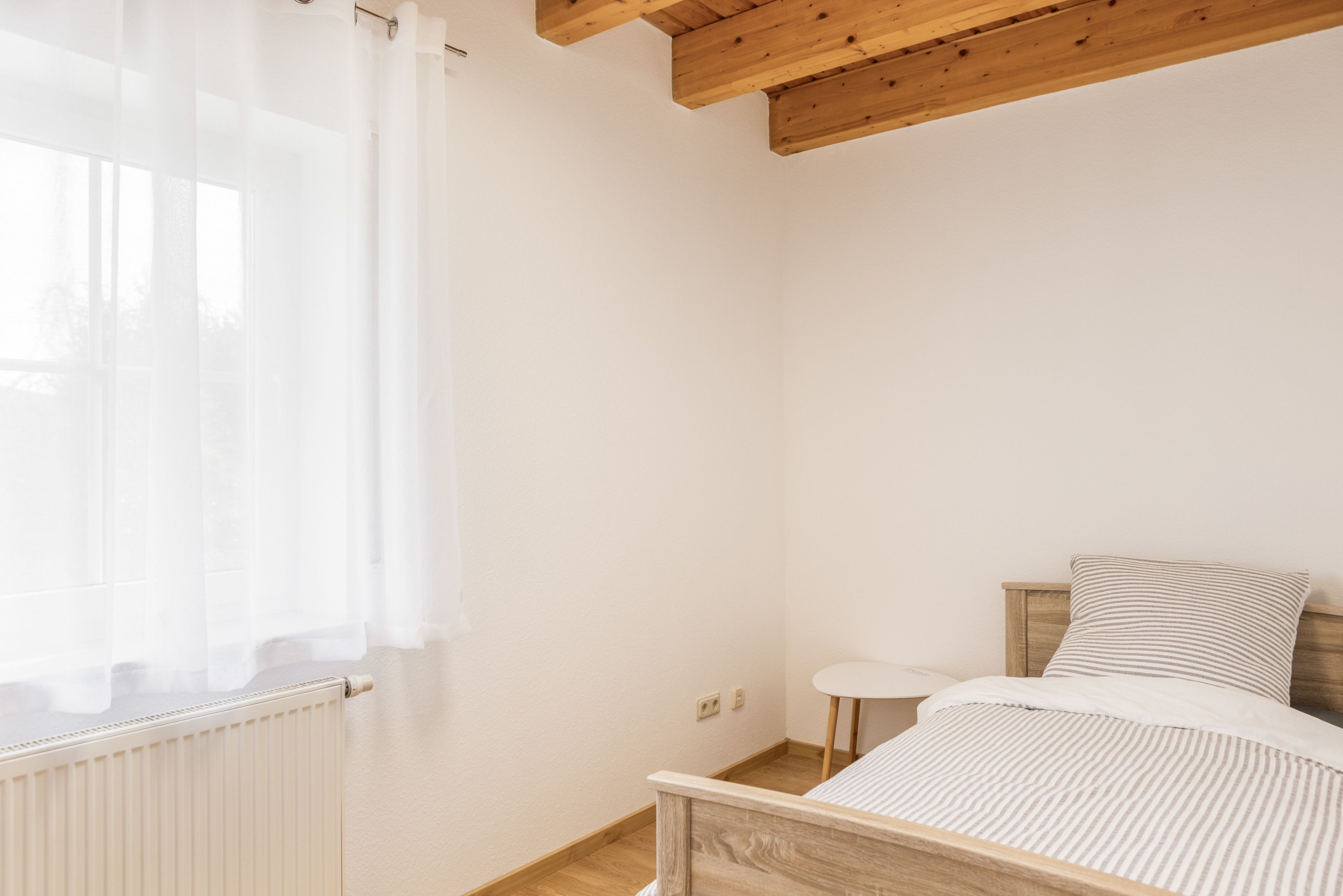 Une chambre à coucher avec une tête de lit en bois, un lit simple, une petite table de chevet et une fenêtre ornée de rideaux légers.