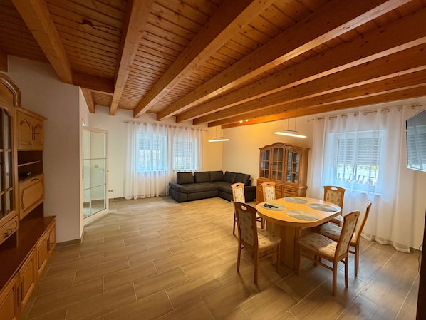 Een ruime woonkamer met houten plafond, een bank, een eettafel met stoelen en een kast.