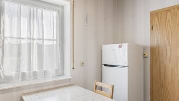 Une petite cuisine équipée d’un réfrigérateur, d’une fenêtre ornée de rideaux légers et d’une porte en bois.