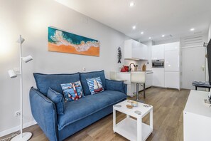 Uma sala de estar moderna com um sofá azul, uma mesa de centro e uma área de cozinha com armários brancos e uma chaleira vermelha.