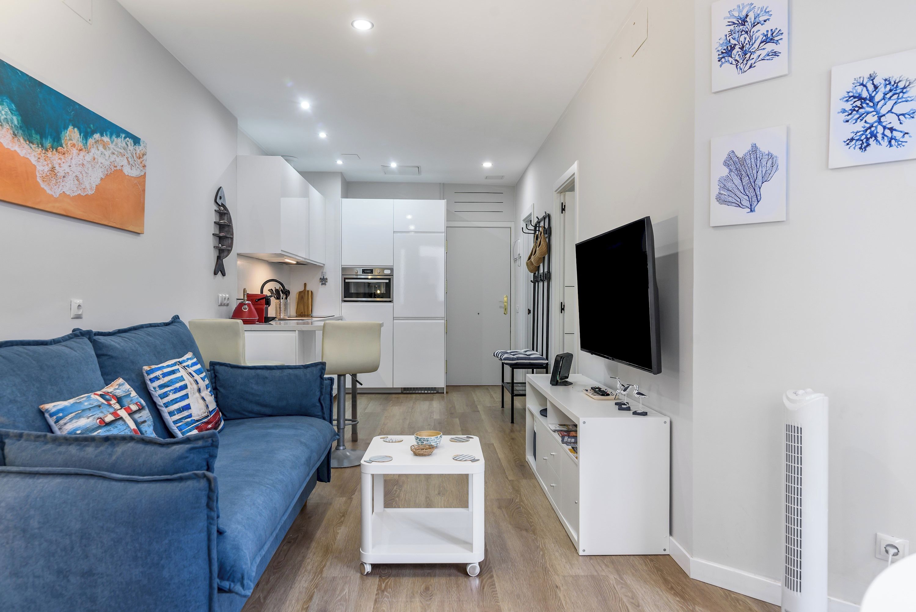 A modern living room with a blue sofa, a coffee table, and a flat-screen TV. A kitchen with white cabinets and a stove is visible in the background.