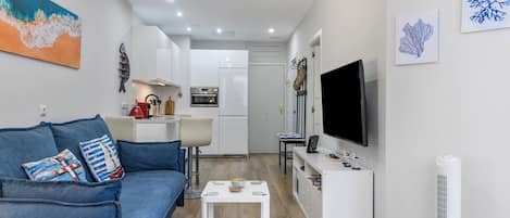 A modern living room with a blue sofa, a coffee table, and a flat-screen TV. A kitchen with white cabinets and a stove is visible in the background.