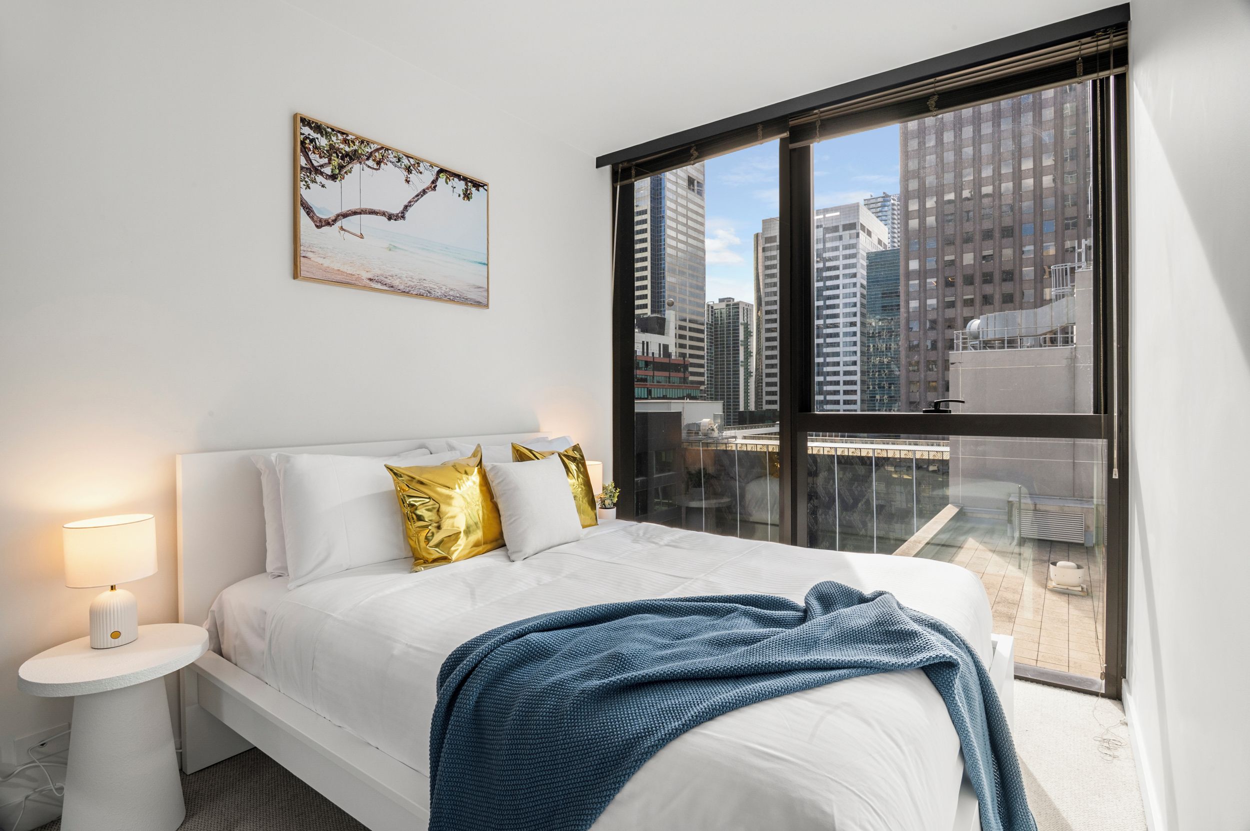 A modern bedroom with a large bed, white bedding, a blue throw blanket, and a city view through a large window.