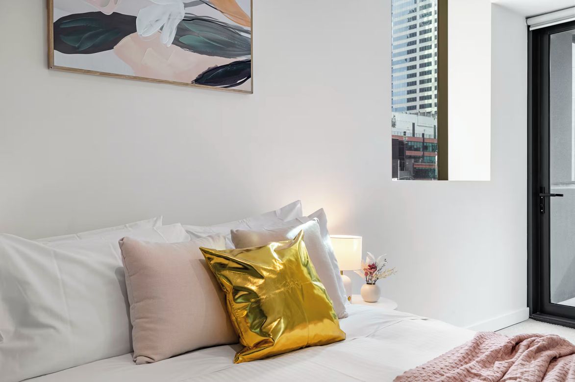 A hotel room with a bed, pillows, a painting, a window, and a view of buildings.