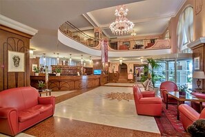 A spacious lobby with a grand chandelier, a reception desk, and a staircase leading to an upper level. - Hotel Astoria (Grado)