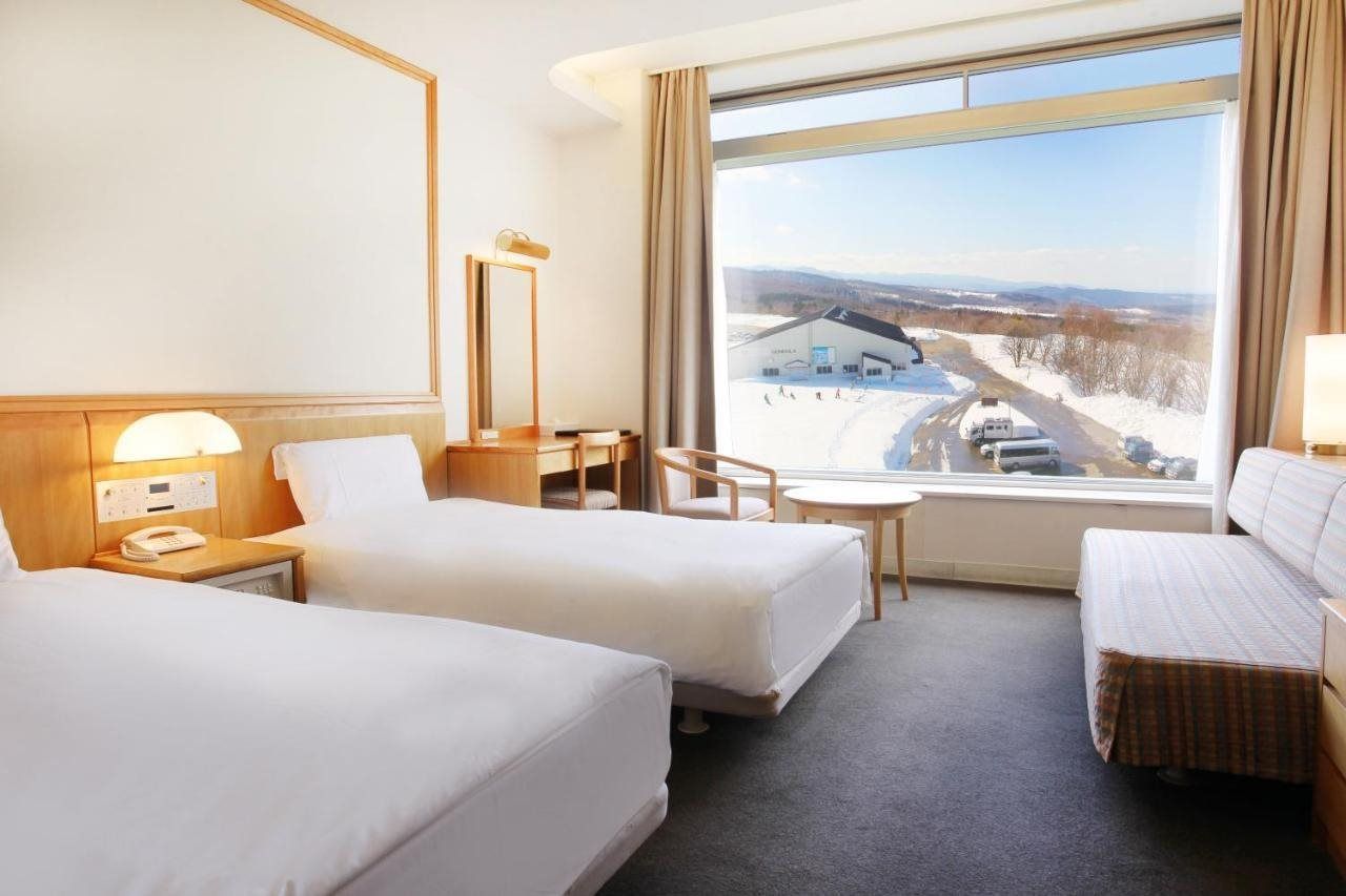 Ein Hotelzimmer mit zwei Betten, einem Schreibtisch, einem Stuhl und einem großen Fenster mit Blick auf eine verschneite Landschaft.
