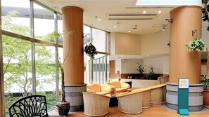 Un hall de hotel con grandes ventanales, una recepción, muebles de mimbre y plantas en macetas.