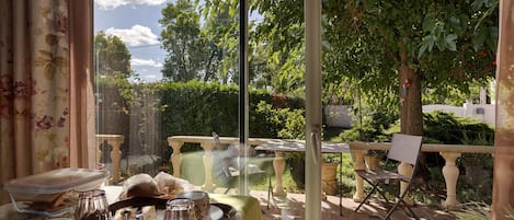 A dining area with a table set for two, a chair, and a view of a garden through a glass door.
