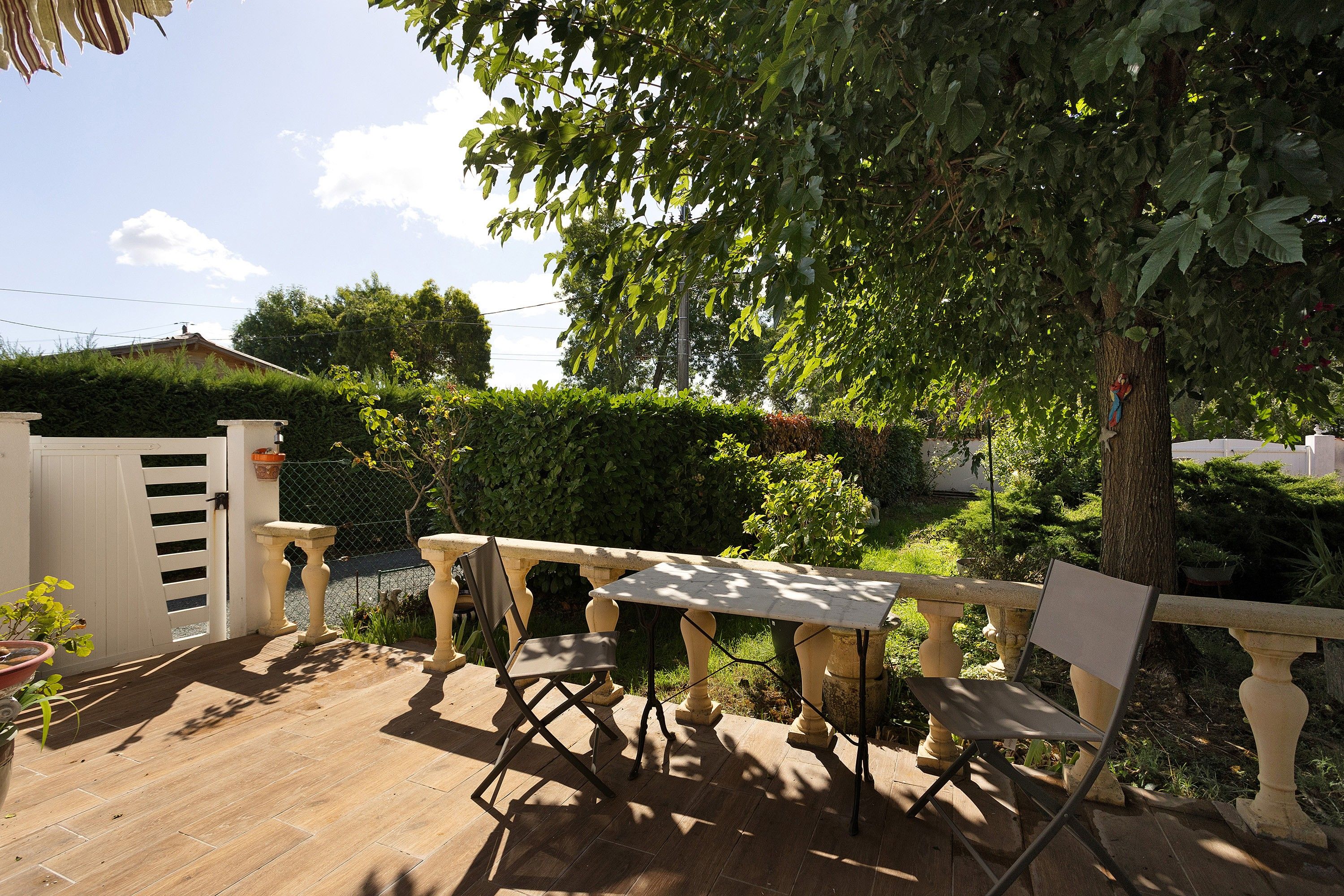 Une terrasse avec une table et des chaises, entourée de verdure et d’une clôture blanche.