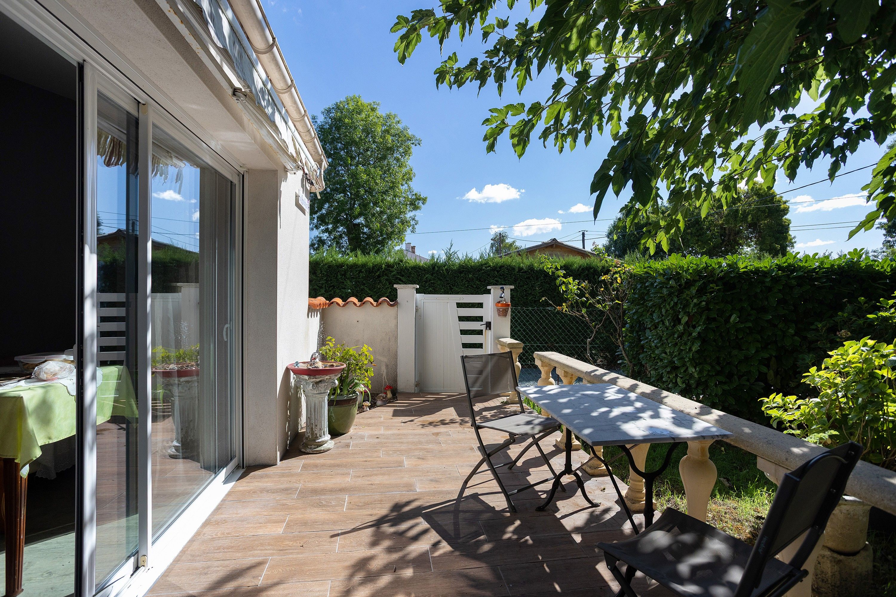 Une terrasse avec une table et des chaises, un pot de fleurs et une vue sur une haie et des arbres.