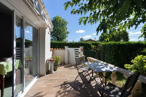 A patio with a table and chairs, a potted plant, and a view of a hedge and trees.