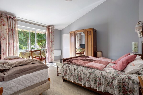A bedroom with two beds, a wooden wardrobe, a table with chairs, and a window with curtains.