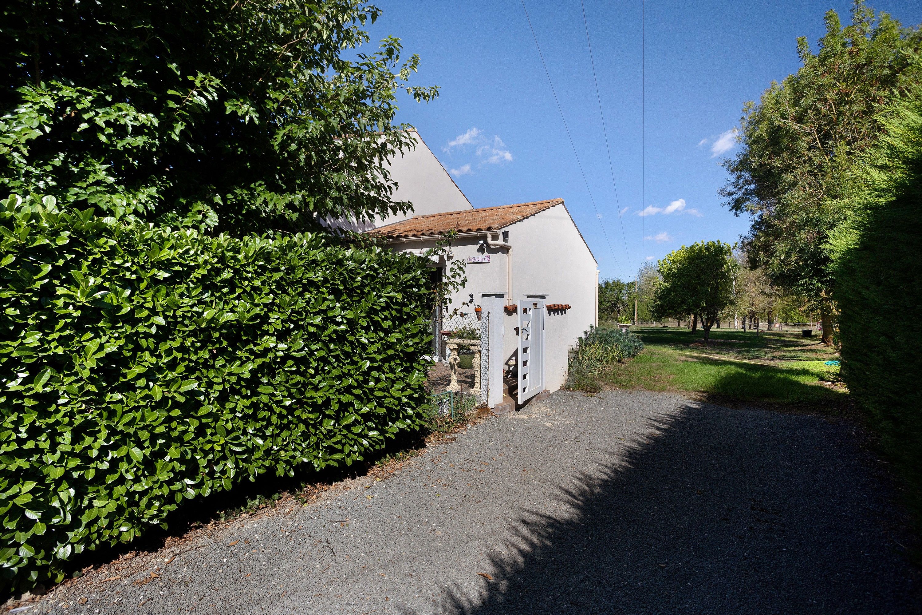 Un bâtiment à la façade blanche et au toit recouvert de tuiles, entouré de verdure et bordé d’un chemin de gravier.