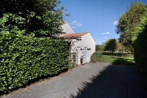 Un bâtiment à la façade blanche et au toit recouvert de tuiles, entouré de verdure et bordé d’un chemin de gravier.