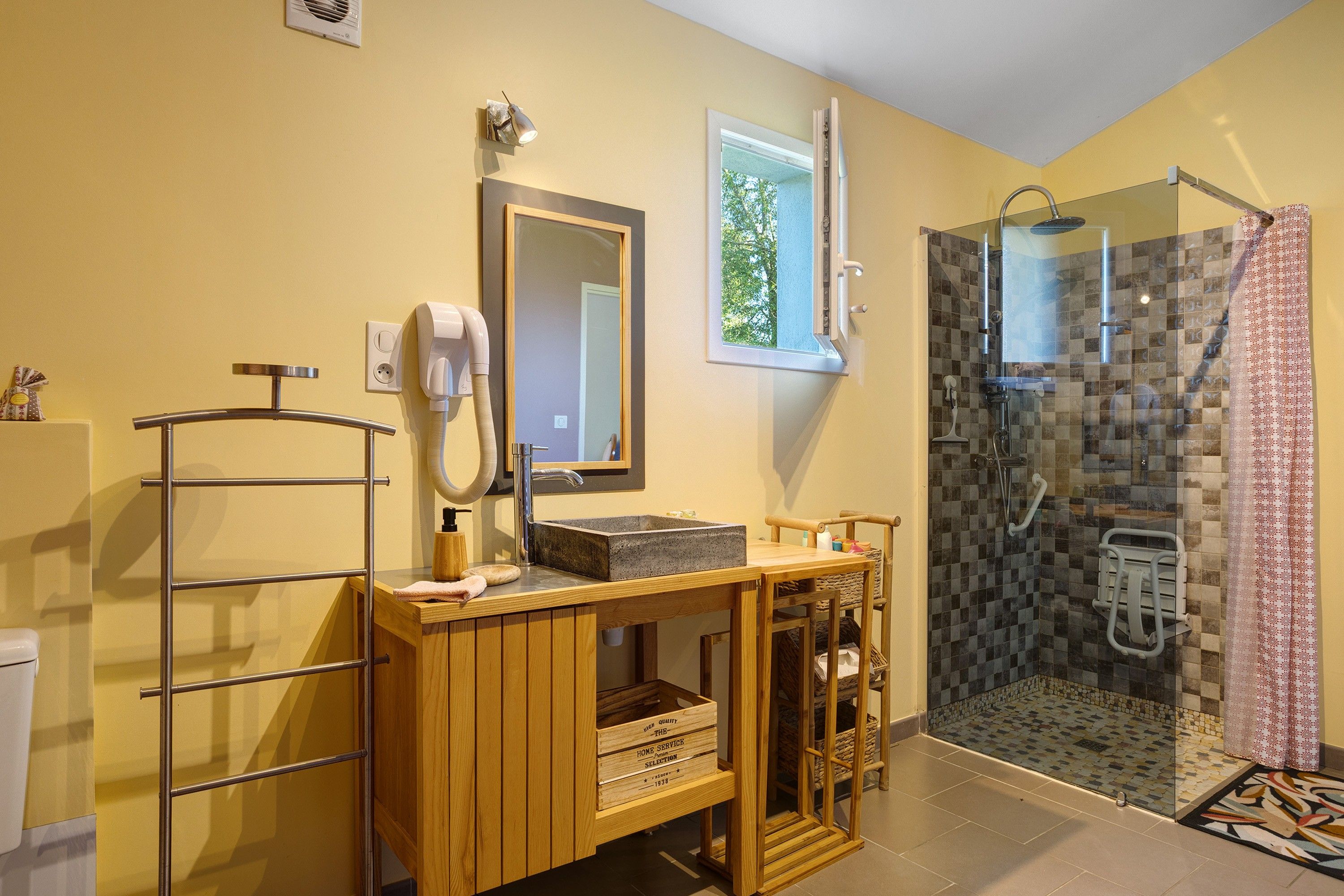 Un baño con tocador de madera, un espejo, un secador de pelo, ducha con puerta de cristal y una ventana pequeña.